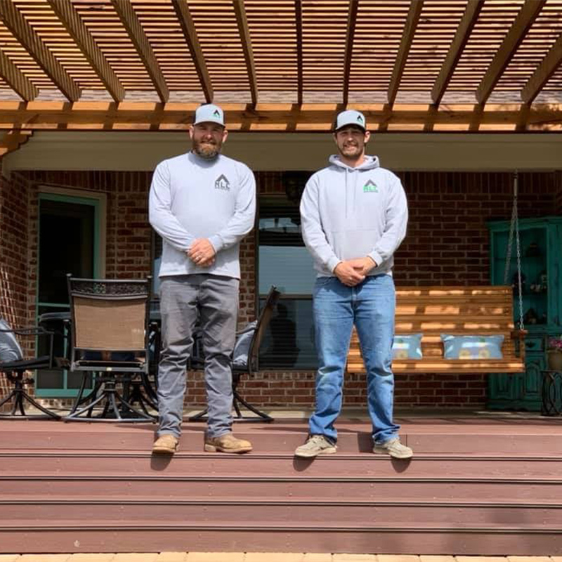 Two roofers are standing on a wooden deck with a wood pergola overhead. They are outside and looking at the camera.