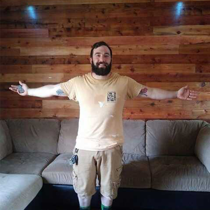 A roofer is standing in front of a couch and his arms are spread out in gratitude.